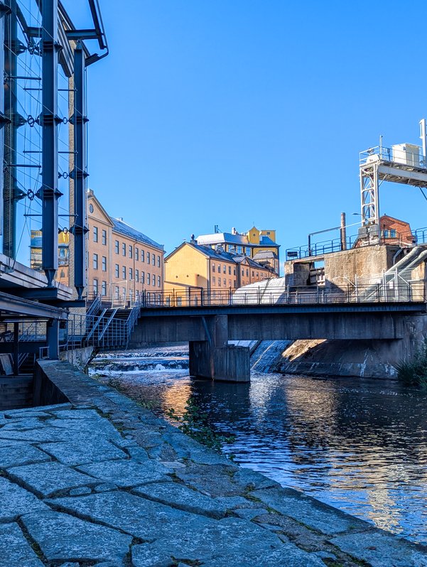 Looking upstream towards the falls from the park outside the lower level of the Louis de Geer Concert & Congress. The blue sky is reflected off the glass windows of the concert hall at the left, and off the river flowing towards the lower right. The falls are partly visible behind a concrete bridge. Rose-gold buildings rise behind them, center, while industrial machinery is still visible on the upper right.