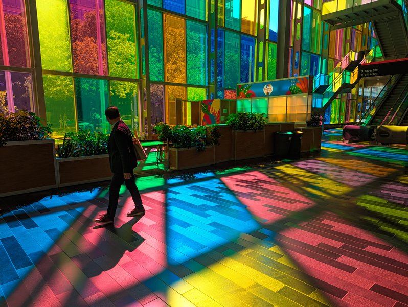 In the west foyer of the Palais des Congrès de Montréal, sunlight streams through a wall of rectangular stained glass panels, tiling the floor with trapezoids of color. Through the panels can be seen the trees of Jean-Paul-Riopelle Place, people among the trees, and more buildings behind the trees. A man walks through the foreground in business casual clothes, a tote bag over his shoulder, face turned towards the stained glass wall behind him; his shoes, shoulders, and the top of his head are colored red like the floor below him.